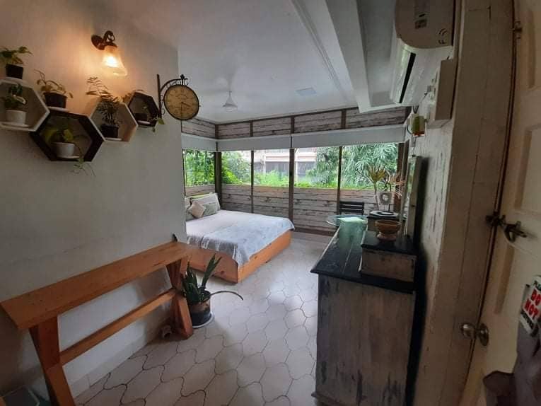 A bright and airy room is presented with large glass windows opening to a view of lush greenery. The space features a spacious bed and a rustic wooden table, paired with lively potted plants. Air conditioning and a wall clock contribute to a comfortable environment.