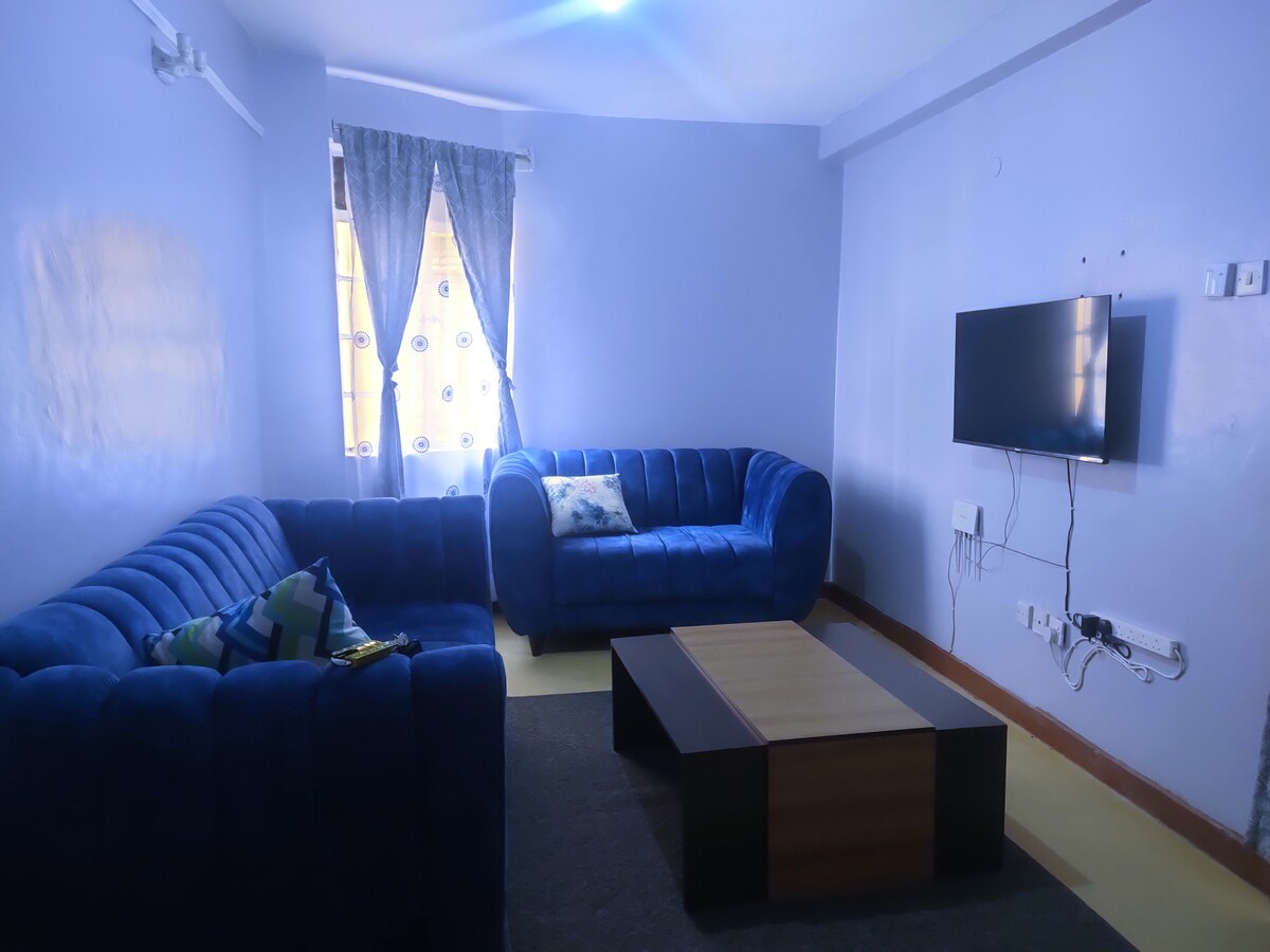 A cozy living area is presented, featuring a pair of blue sofas arranged around a coffee table. A wall-mounted television is visible opposite the seating. Natural light comes through a window adorned with sheer curtains, adding brightness to the room.