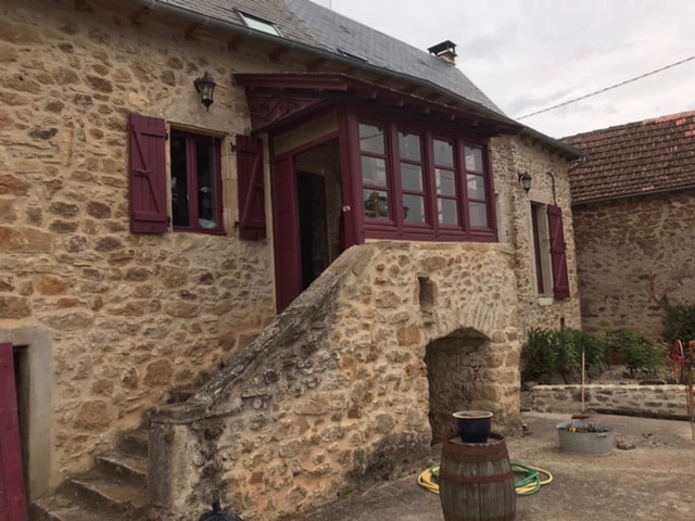 A rustic stone building is featured, showcasing deep maroon shutters and a series of large windows that allow natural light to illuminate the interior. The entrance is marked by a stone staircase leading up to the front door, adding a charming touch to the inviting exterior.