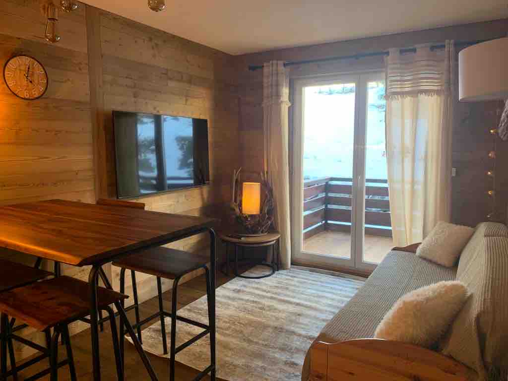 A living area features wooden walls and a textured rug, complemented by a wall-mounted television. Glass doors provide access to a balcony, allowing natural light to enter. A wooden dining table with tall chairs is positioned beside a cozy sofa.