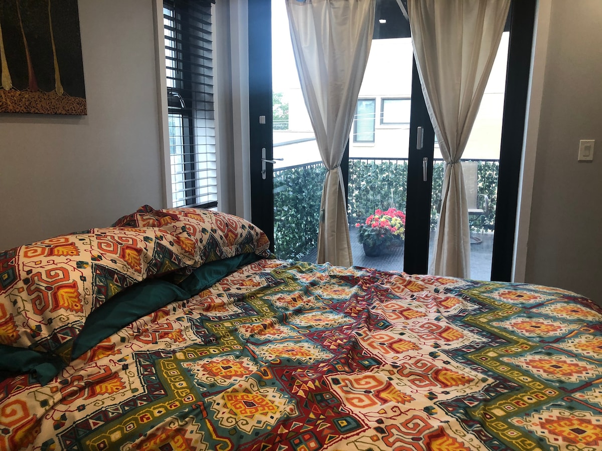 A spacious bed layered with a colorful patterned blanket is positioned near large glass doors that open to a private deck. Sheer curtains frame the entrance, allowing natural light to fill the room while offering a view of the greenery and flowers on the deck.