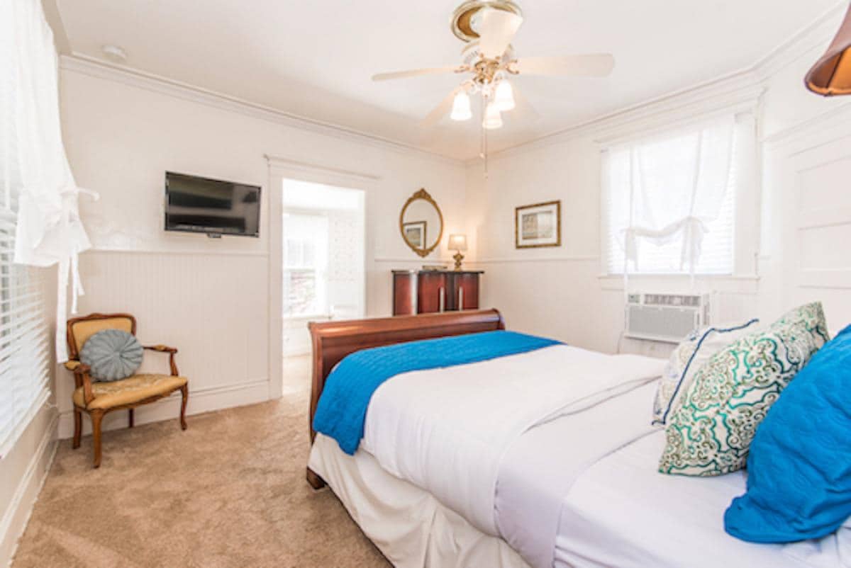 A bright bedroom is depicted with a spacious bed dressed in white linens and an accent blanket in turquoise. A vintage armchair is positioned beside the bed. Light-colored walls and large windows allow natural light to enhance the space, which features modern amenities like a mounted television.