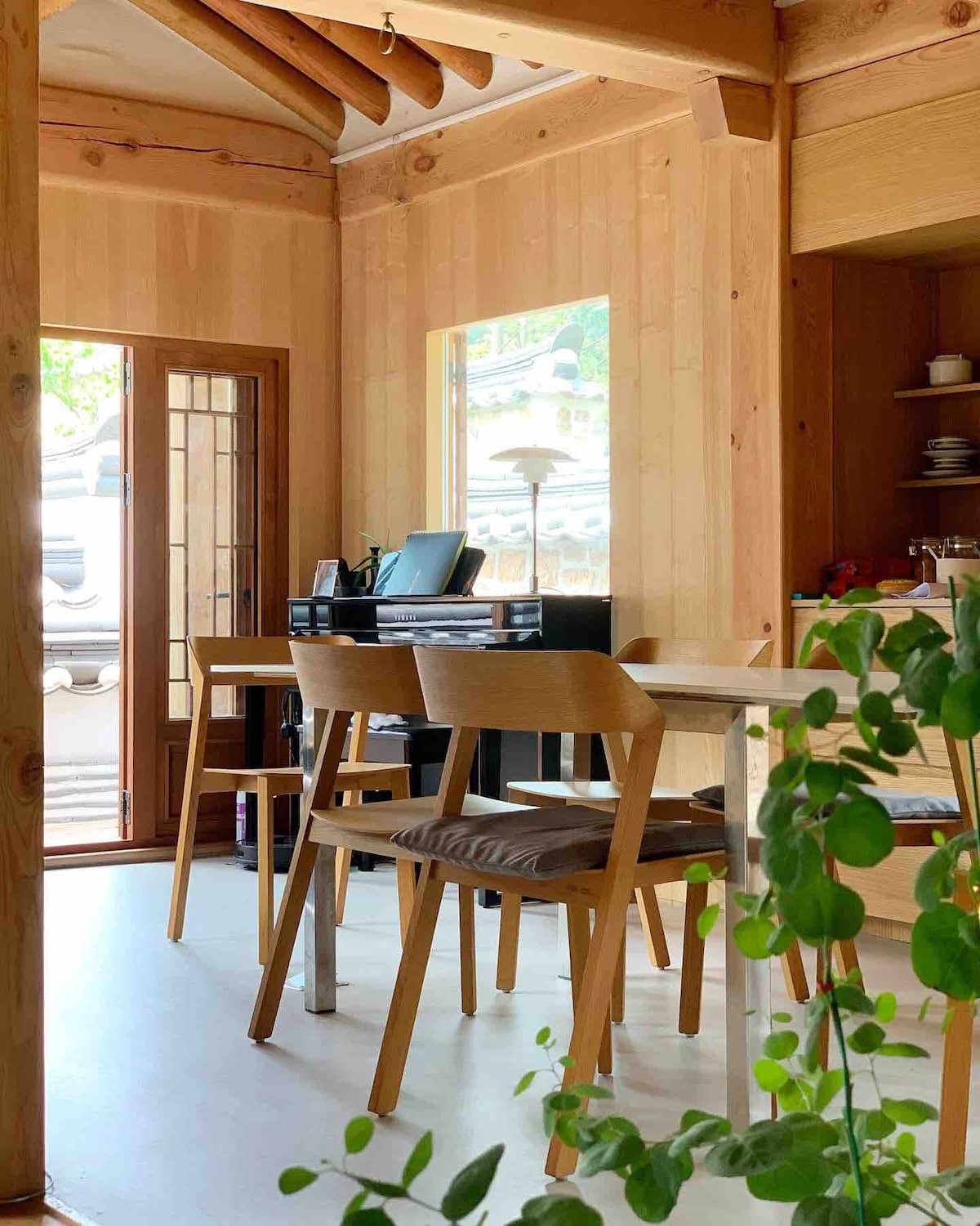 A wooden dining area features a table complemented by four chairs, creating a welcoming space for gatherings. Bright natural light streams through a large window, illuminating the room's warm wooden paneling. A glimpse of greenery adds a touch of nature to the interior ambiance.