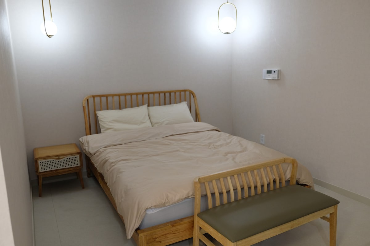 A sleeping area features a wooden bed with a light-colored comforter. Two soft pillows rest on the bed, with a wooden bench positioned at its foot. Soft lighting is provided by two wall-mounted fixtures, enhancing the calm color scheme of the room.