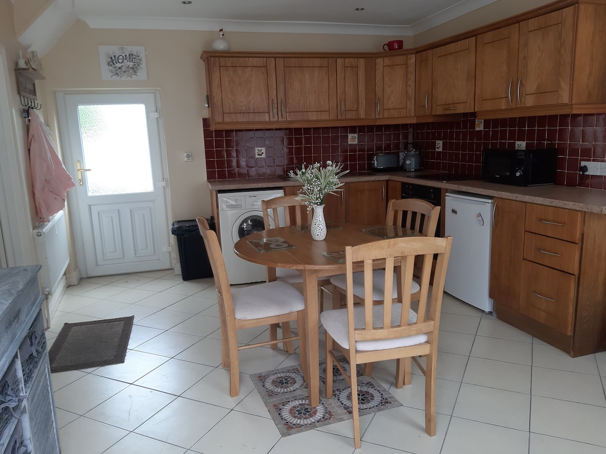 The bright kitchen area features wooden cabinetry and a round dining table surrounded by four wooden chairs. Appliances, including a washing machine and refrigerator, are visible. A door leads outside, and a small table decoration adds a touch of charm.