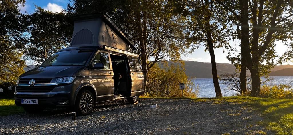 A modern VW campervan is parked on a gravel area near a serene body of water. Trees frame the scene, and sunlight filters through their leaves, casting gentle shadows. The van's side door is open, inviting a sense of exploration in nature.