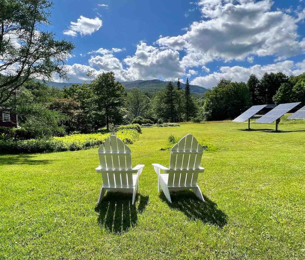 Two classic white Adirondack chairs are positioned on a spacious green lawn, facing a scenic view of rolling mountains under a bright blue sky. Solar panels stand to the right, surrounded by trees and lush foliage.