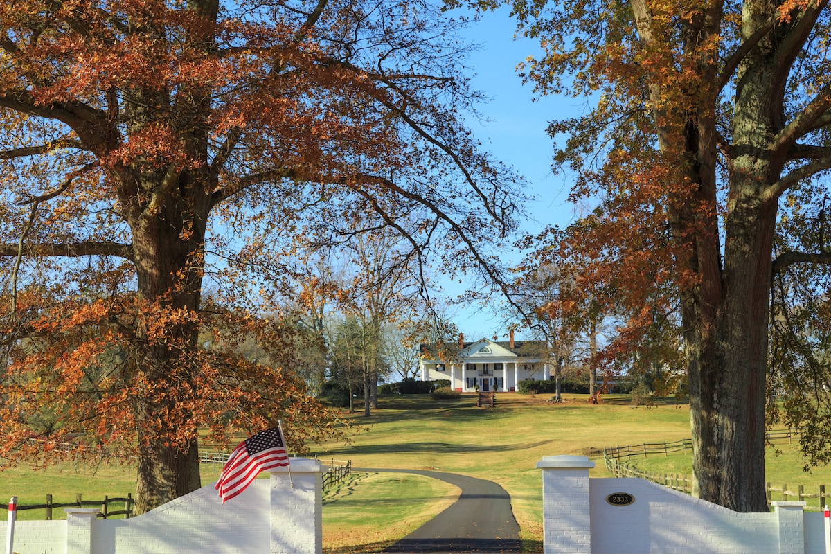 A scenic entryway is framed by large trees displaying vibrant autumn foliage. A white entrance gate, complete with an American flag, leads down a winding driveway toward a stately white mansion set against a backdrop of rolling green lawns.
