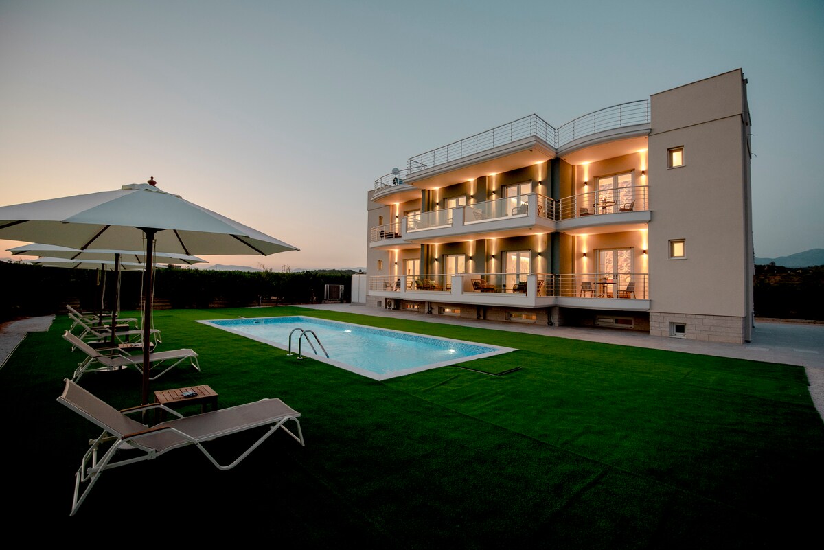 A modern three-story building is depicted, featuring multiple balconies and illuminated interior spaces. A clear blue pool is situated in front, surrounded by lounge chairs and umbrellas on manicured grass. The evening sky casts a soft glow over the serene outdoor setting.