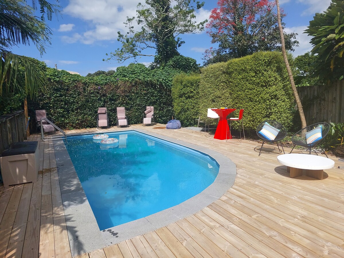 A serene outdoor space is shown, featuring a clear pool surrounded by wooden decking. Lounge chairs are positioned beside the water, and a bright red table with matching chairs invites gathering. Lush greenery encircles the area, enhancing the tranquil atmosphere.