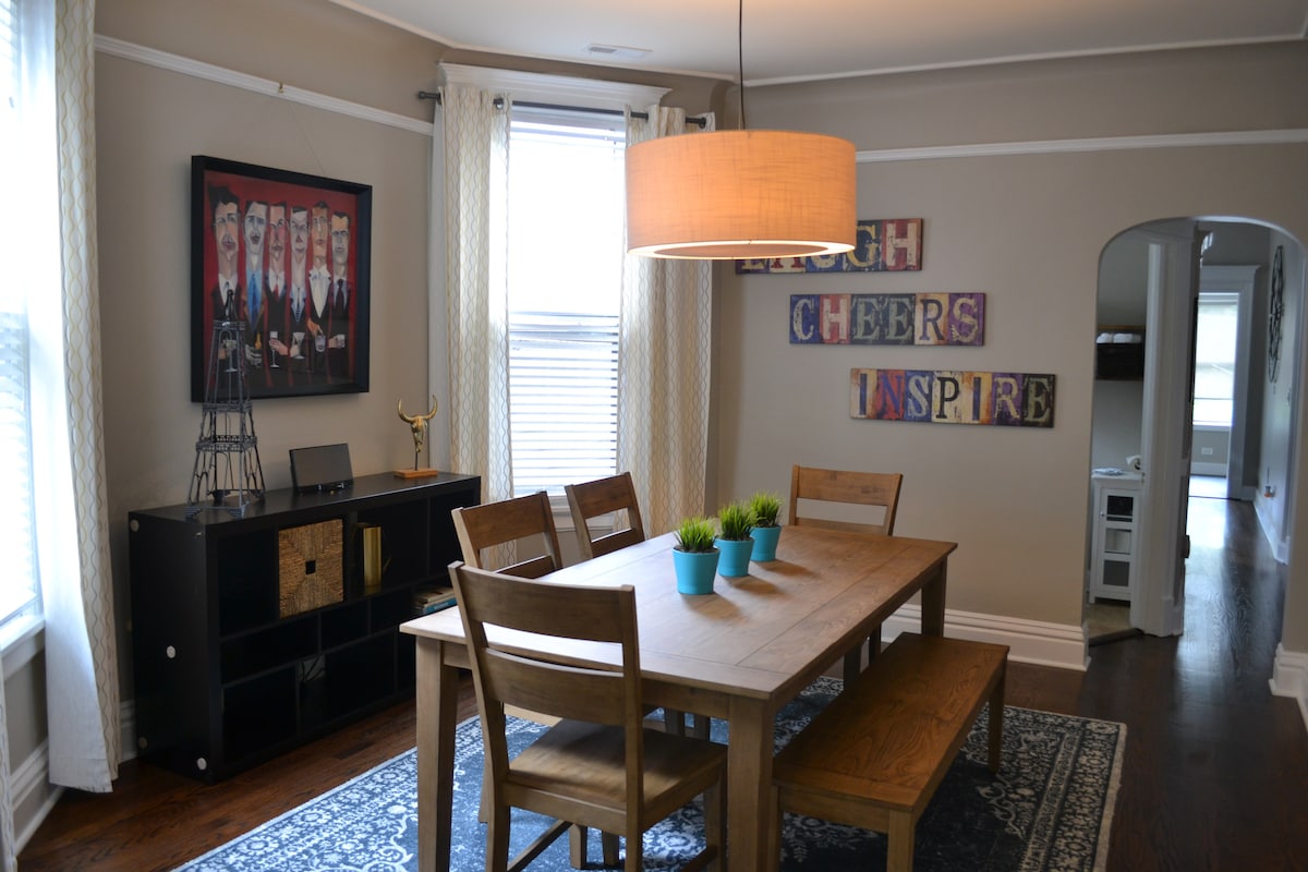 An inviting dining area features a wooden table surrounded by four chairs. A large pendant light hangs above, casting a warm glow. On the table, small potted plants add a touch of greenery. A wall displays framed artwork, and natural light filters through window treatments.
