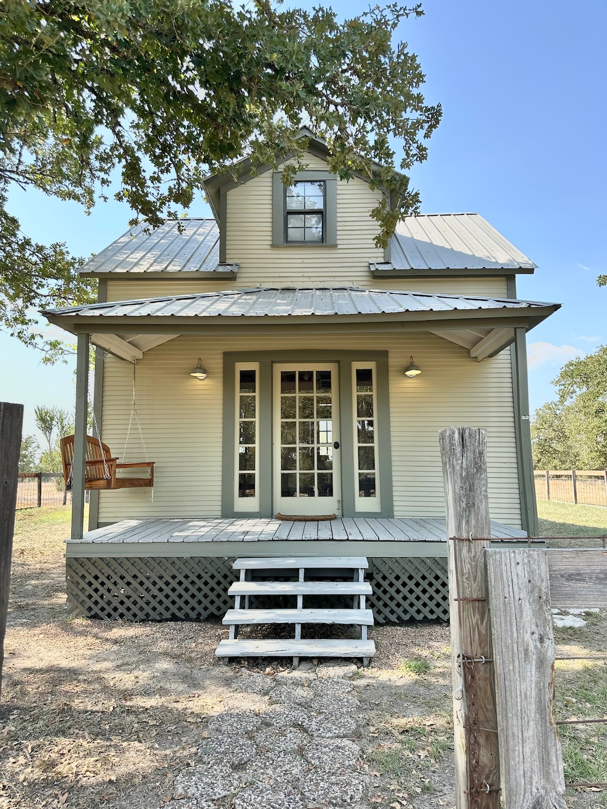 The exterior of the cottage features a welcoming front porch with a swing and a charming double-door entrance. A metal roof and light green siding complement the rustic surroundings. Steps lead up to the porch, while a fence and grassy area create an inviting atmosphere.