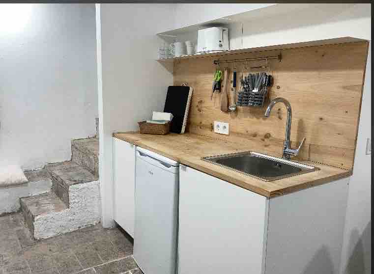 A compact kitchen area features a countertop made of wood and includes a sink with a modern faucet. Below, a refrigerator is positioned next to a white storage unit. Hanging utensils and cooking tools are visible on a wall-mounted rack. Stairs lead to another level.