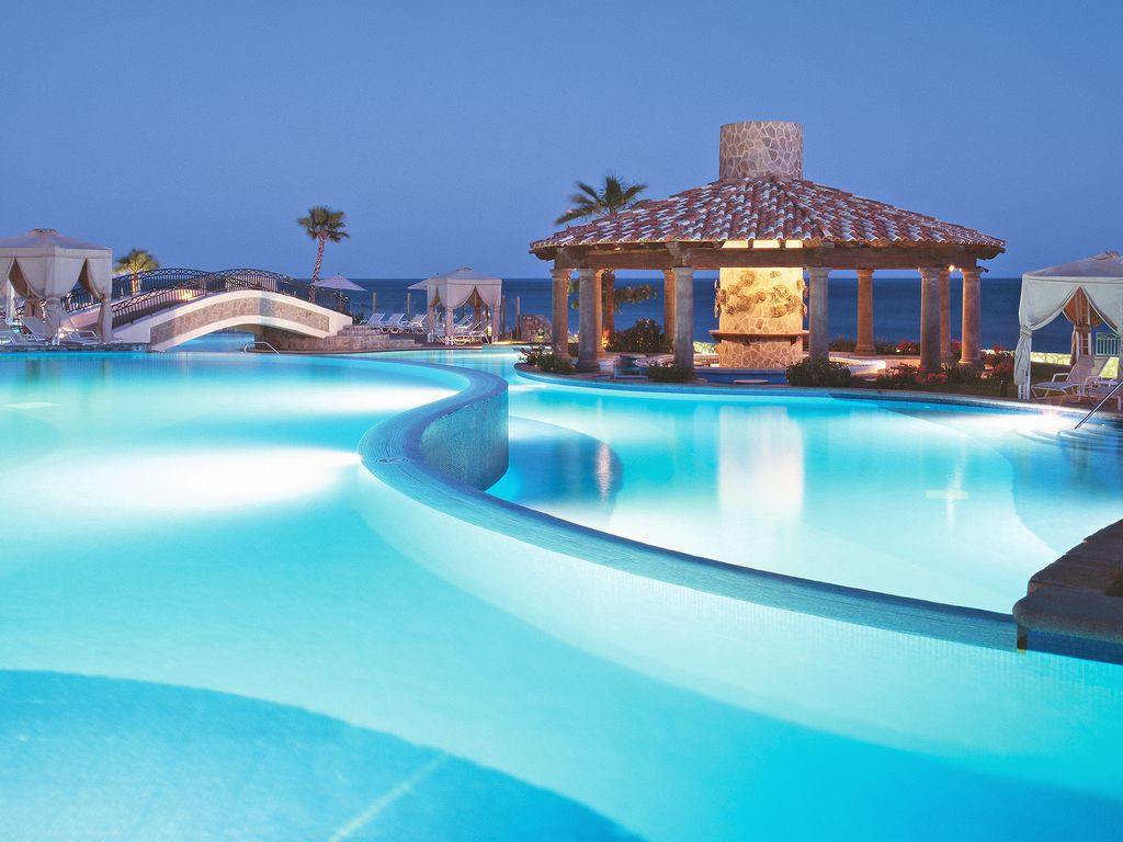 A tranquil pool area is highlighted by a gently curving design, featuring illuminated water reflecting the evening sky. A stone gazebo offers shaded seating, while palm trees stand in the background, enhancing the serene beachfront ambiance.