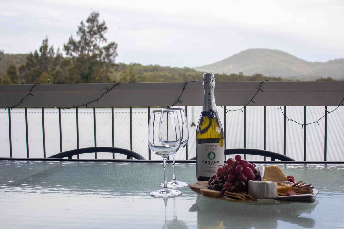 A glass table is set outdoors, featuring a bottle of organic sparkling wine alongside two glasses. A wooden platter displays an assortment of cheeses and fresh grapes, with a scenic mountain view in the background.