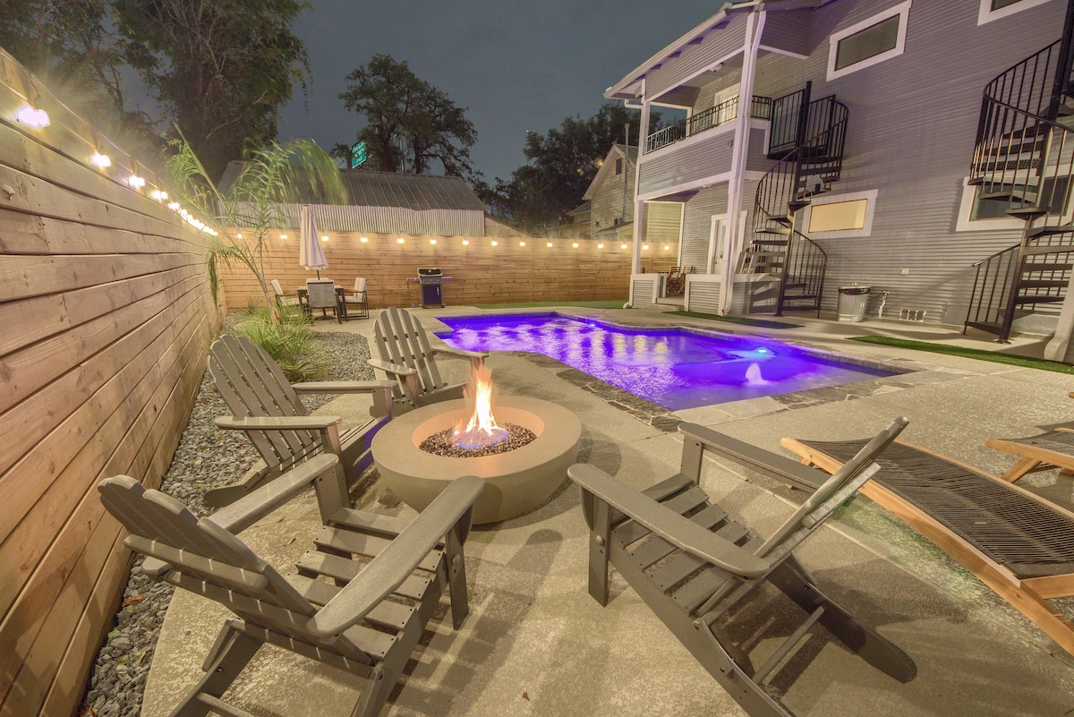 An outdoor space features a circular fire pit surrounded by several wooden adirondack chairs. A swimming pool glows in a calming blue shade, with soft lighting strung overhead adding warmth to the area. Lush greenery is present along the perimeter.