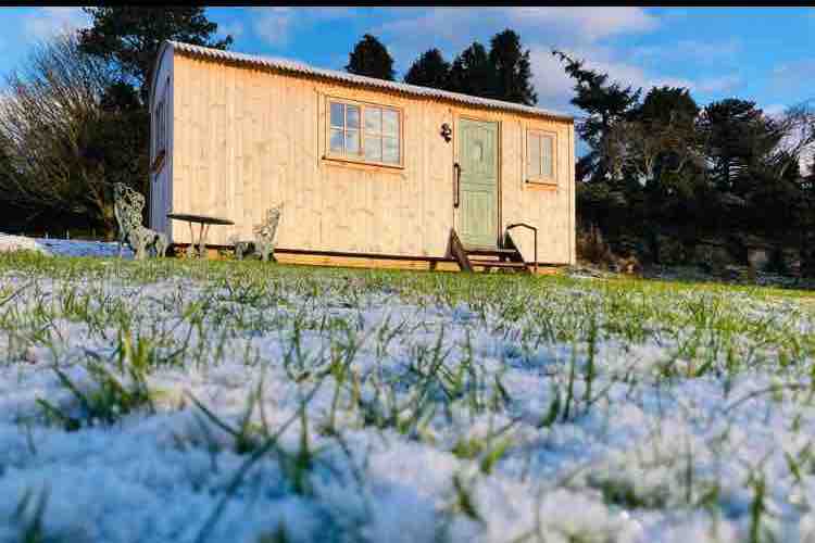 A charming shepherd's hut is set against a backdrop of snow-covered grass. The wooden exterior features a green door and two windows. A small seating area with two chairs is positioned nearby, enhancing the peaceful rural atmosphere.
