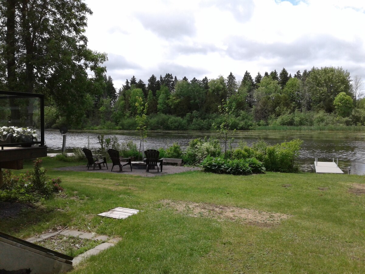 A grassy area borders the water, where several dark wood Adirondack chairs are arranged on a patio. Lush greenery lines the riverbank, adding to the natural setting. A small dock is visible, extending into the calm water, inviting boat access.