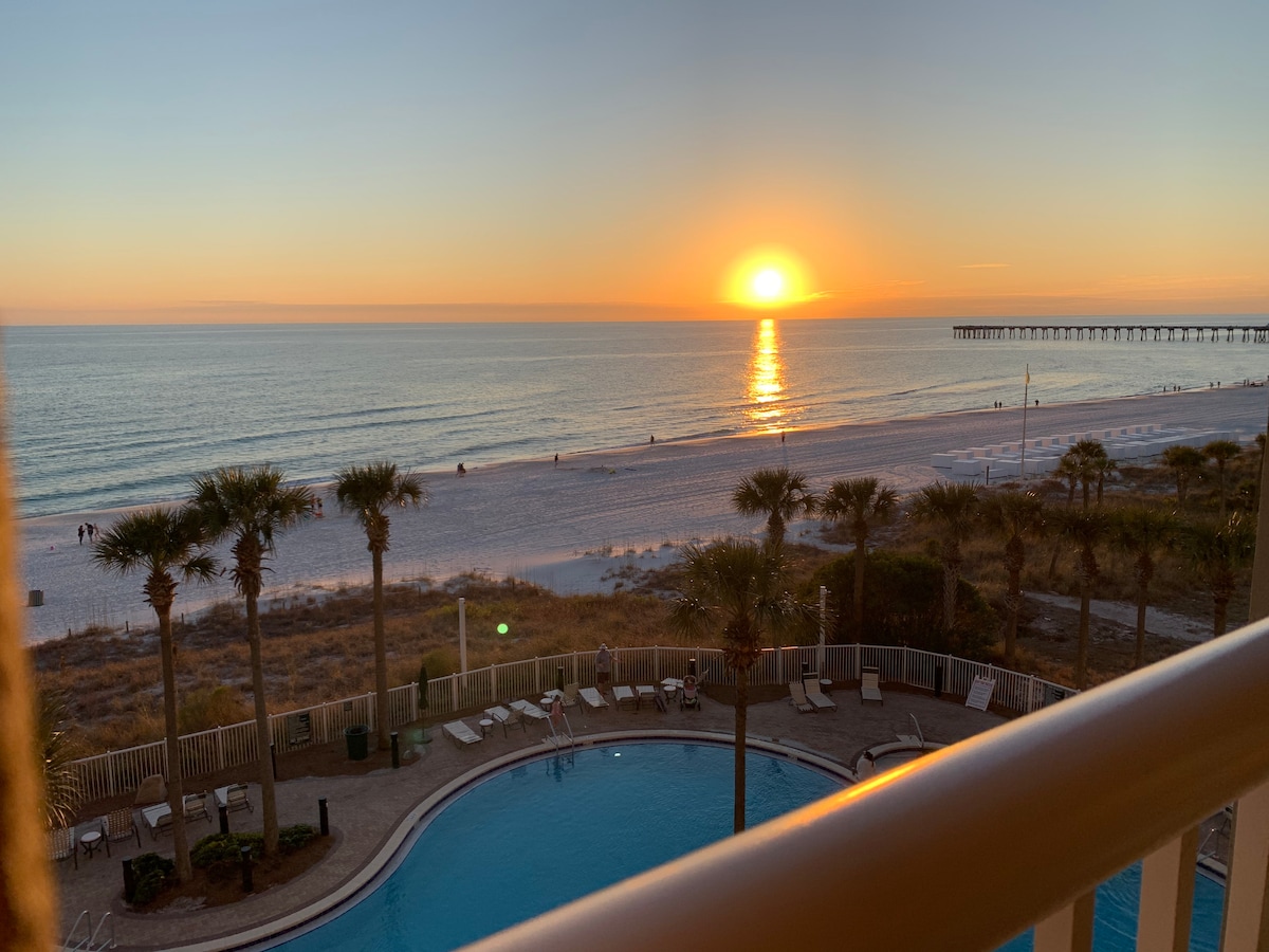 A tranquil beach scene is captured at sunset, with warm hues reflecting on the water. A pool lies in the foreground, surrounded by lounge chairs, while palm trees provide a serene backdrop to the sandy beach stretching toward the horizon.