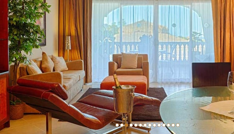 A cozy living space features a large sectional sofa paired with an orange accent chair. A stylish chair is positioned nearby, with a small table displaying a silver ice bucket. Sheer curtains frame large glass doors that open onto a balcony, allowing natural light to fill the room.