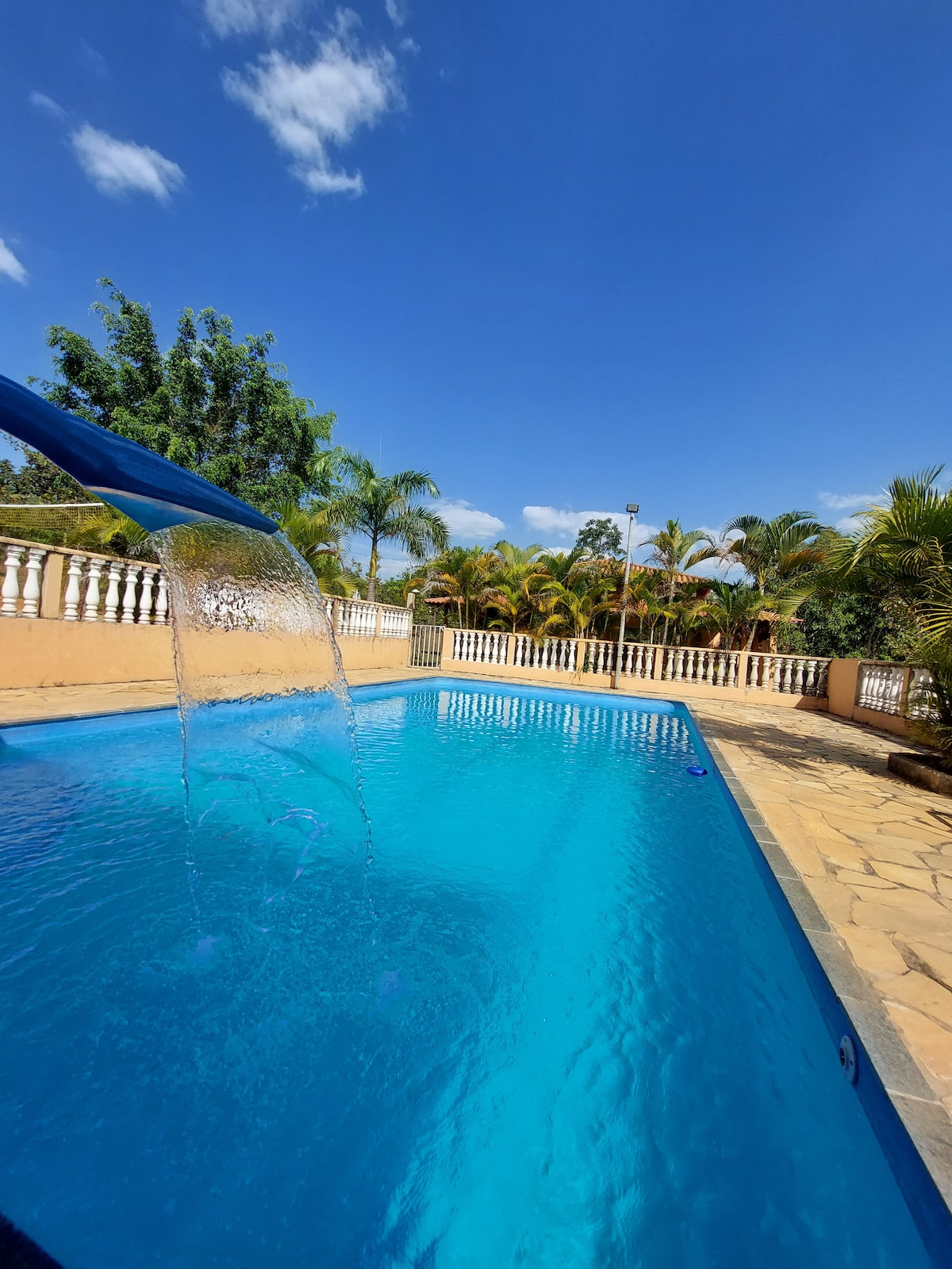 A large swimming pool is showcased, featuring a waterfall that flows gently into the water. Surrounding the pool are palm trees and green foliage under a clear blue sky, creating an inviting outdoor space. The patio area includes stone flooring and a white railing.