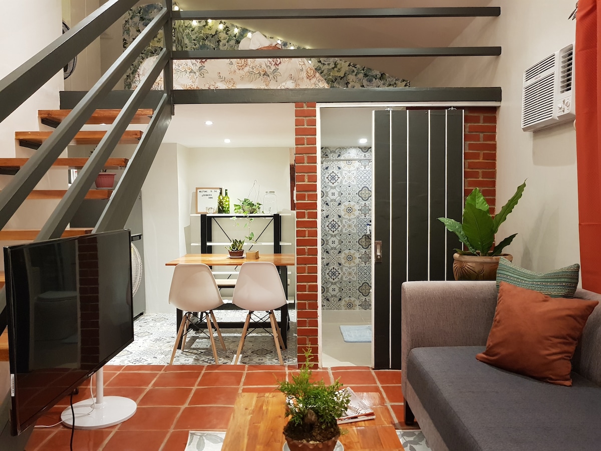 A cozy living area features a grey couch with orange accent pillows, complemented by a wooden coffee table with greenery. A dining table with modern chairs is visible, alongside an air conditioner. The space is characterized by exposed brick walls and a decorative staircase leading to the loft.