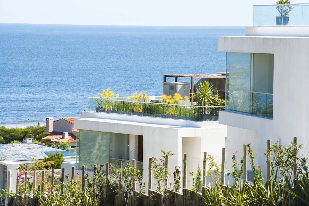 A modern building design features large glass windows and a spacious balcony with greenery. The structure overlooks the ocean, with a clear blue sea visible in the background, enhancing the coastal appeal of the setting.