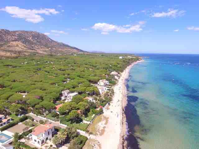An aerial view captures a serene coastal landscape, featuring a sandy beach lined with vibrant greenery. The water displays varying shades of blue, while a gentle shoreline is dotted with beachgoers enjoying the scenery. The surrounding hills contribute to a peaceful atmosphere.