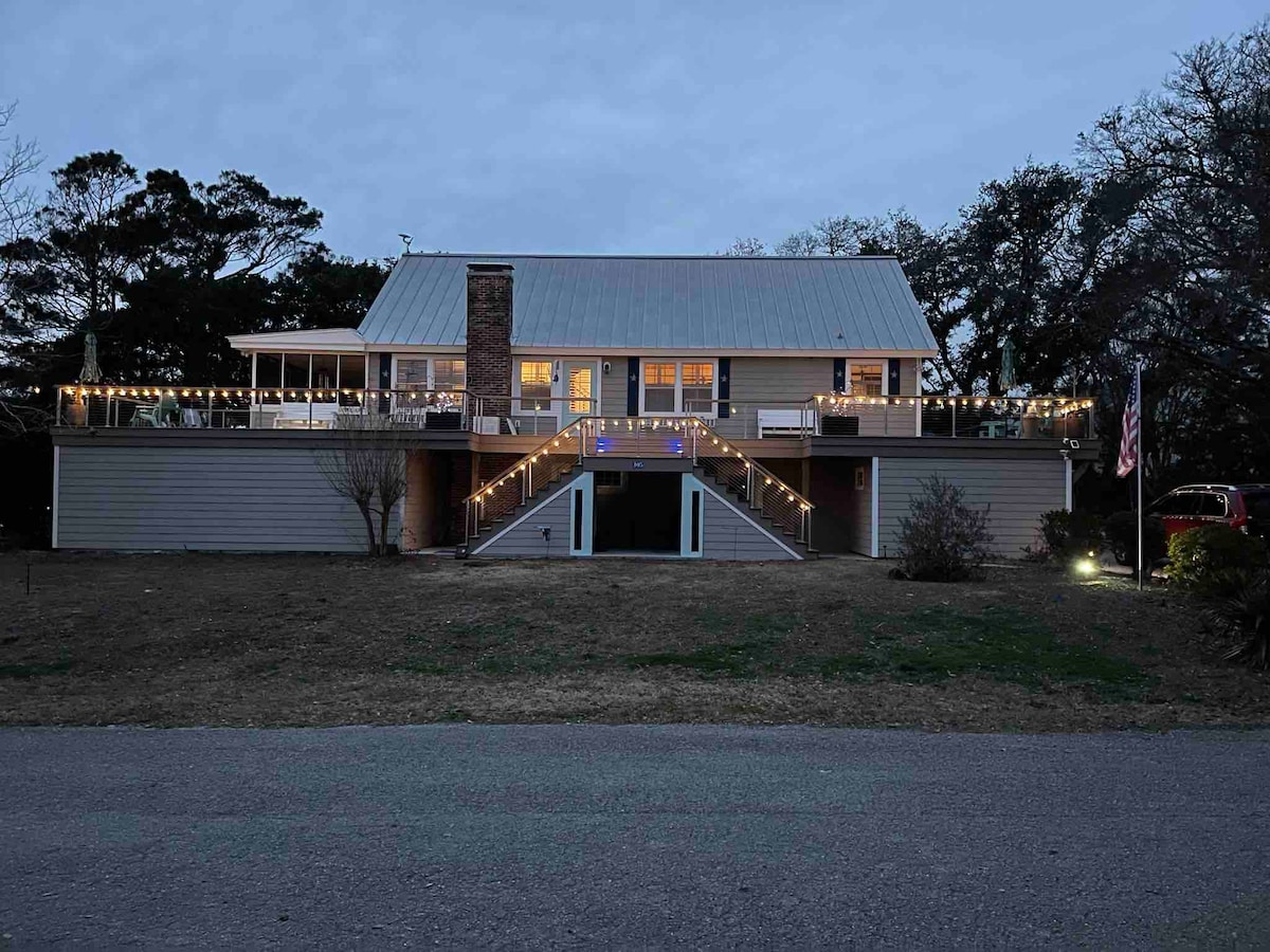 The exterior of a two-story home is illuminated by soft lighting, highlighting the welcoming front porch and steps. A pathway leads to the entrance, flanked by minimal landscaping. An American flag is visible on one side, contributing to the inviting ambiance of the property.