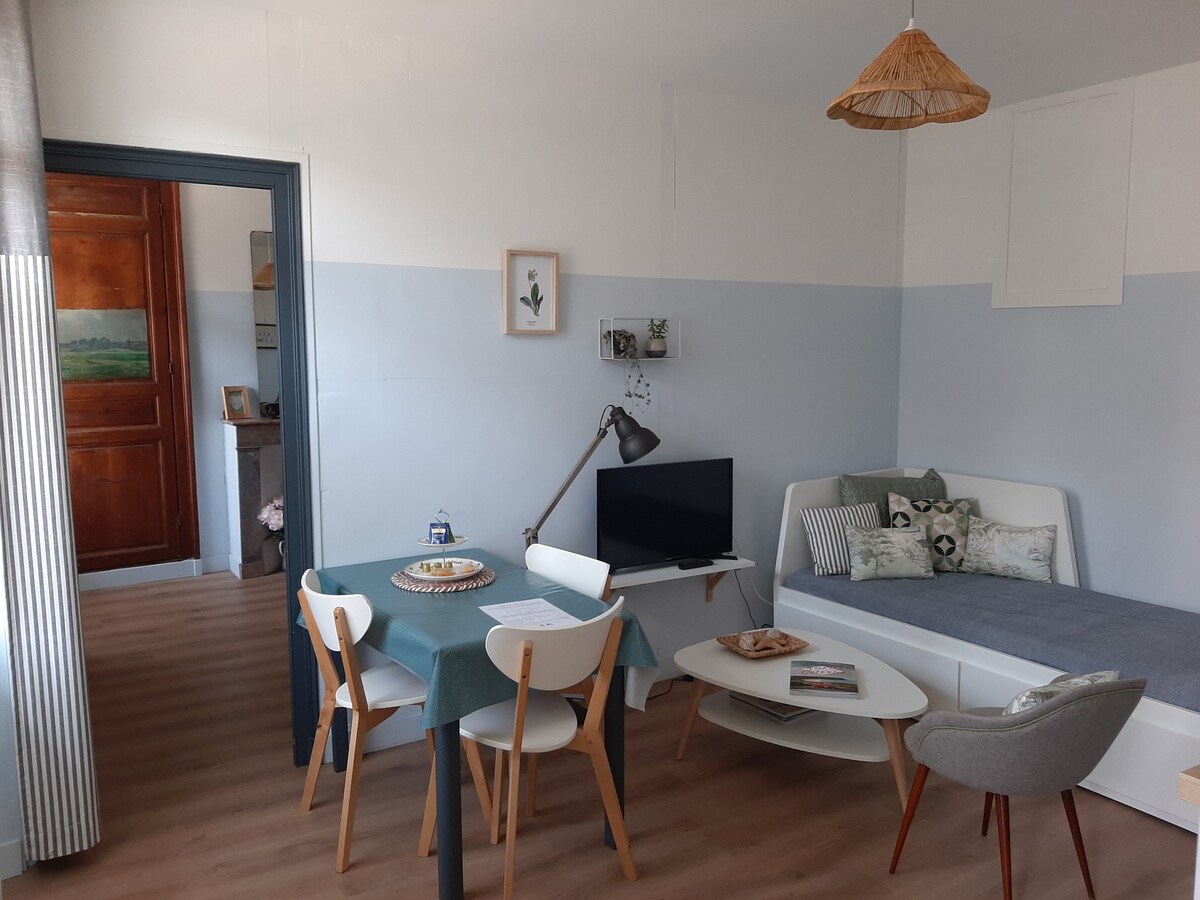 A light-filled living area features a simple dining table set for four, accompanied by a comfortable seating area. A flat-screen TV is positioned next to a modern sofa. The walls are painted in soft blue tones, enhancing the room's fresh ambiance.