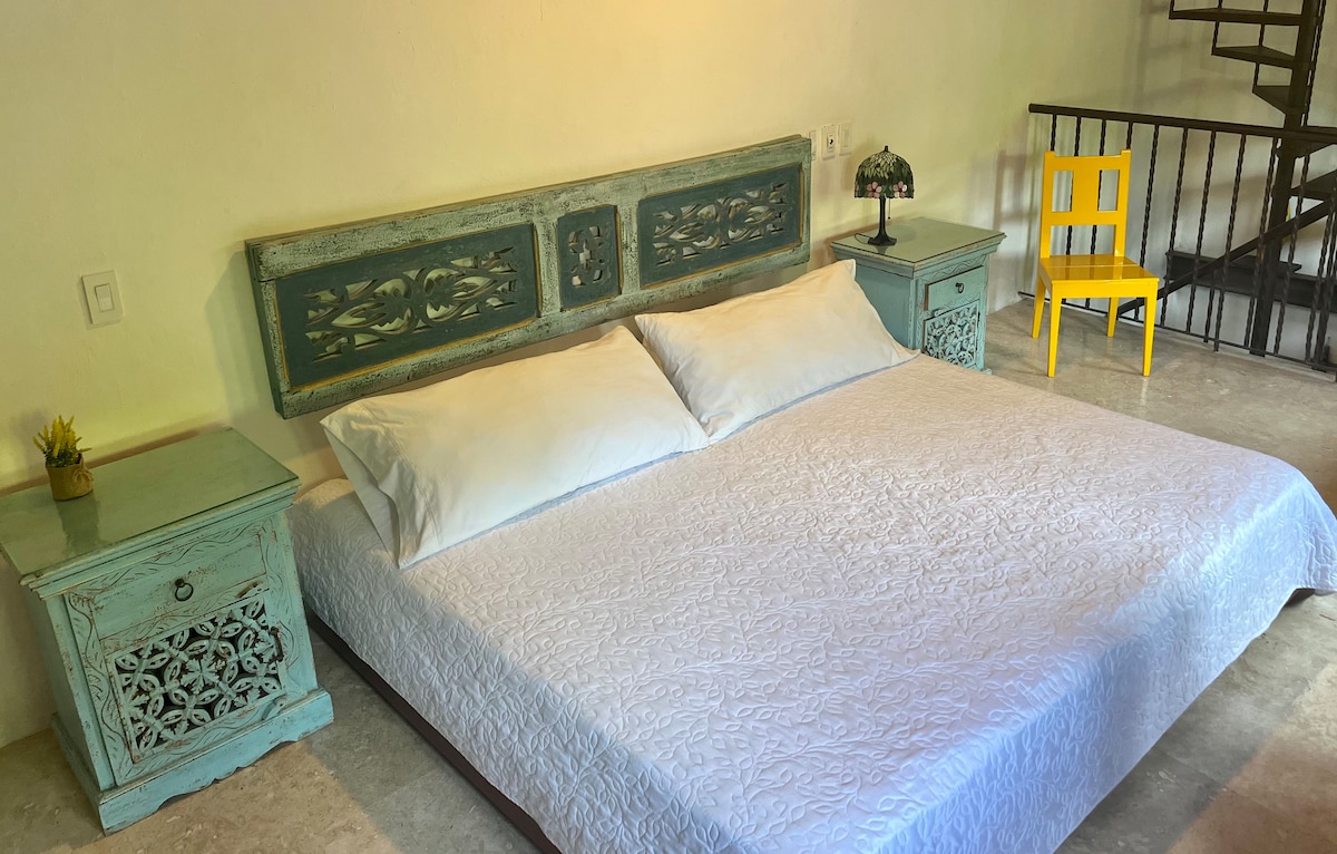 A spacious bed is positioned centrally in the room, adorned with a white quilt. Two light blue nightstands flank the bed, each featuring ornate detailing. A small decorative plant sits atop one nightstand, while a brightly colored yellow chair is present in the corner.