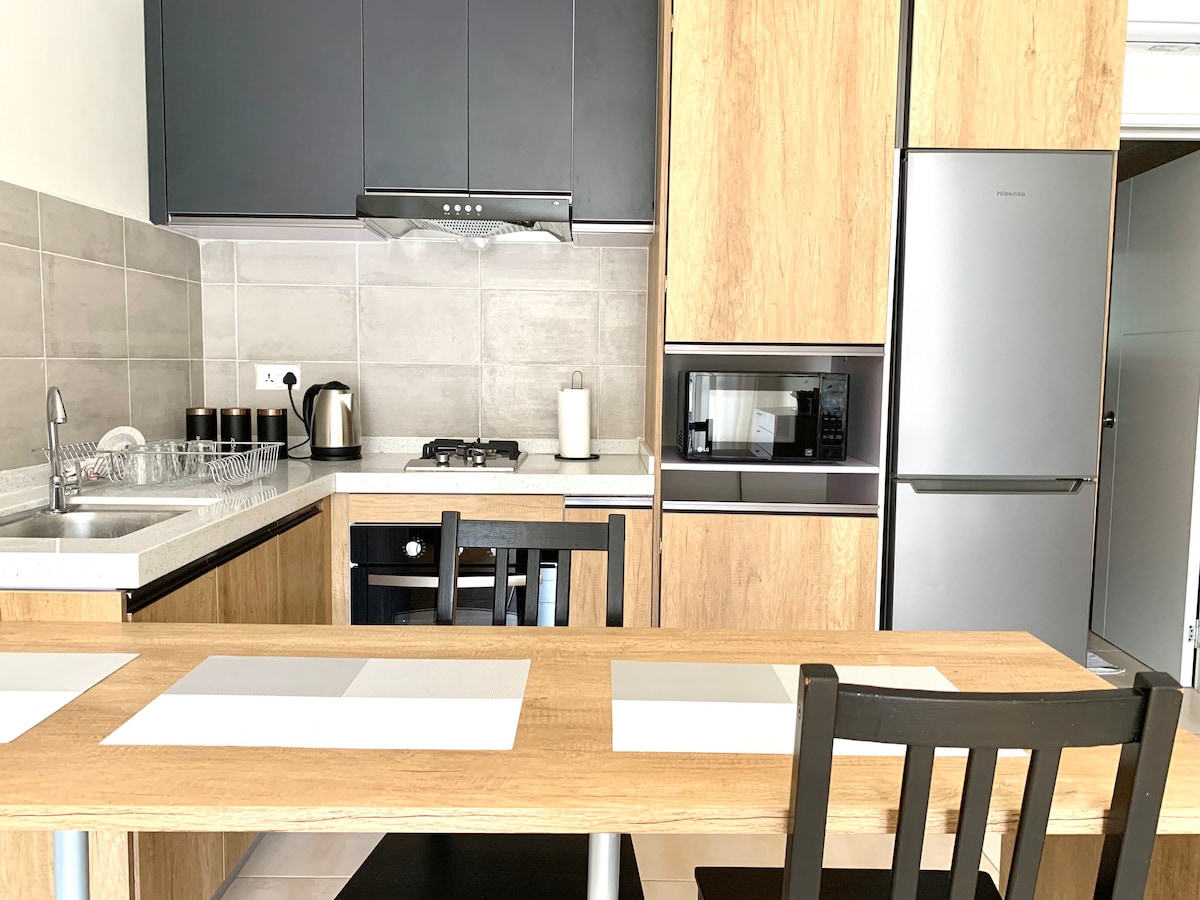 The kitchen features modern cabinetry with a stylish wooden finish. An equipped countertop includes a stove, kettle, and sink, with a microwave and refrigerator integrated into the design. A dining table with black chairs provides space for meals or gatherings.