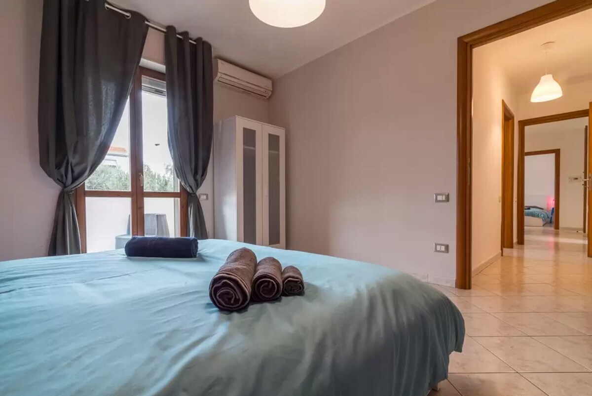 A well-appointed bedroom features a spacious bed dressed in light blue linens. Soft, dark curtains frame a large window that brings in natural light. Rolled towels are neatly arranged on the bed, while a wardrobe and doorways to additional rooms are visible in the background.