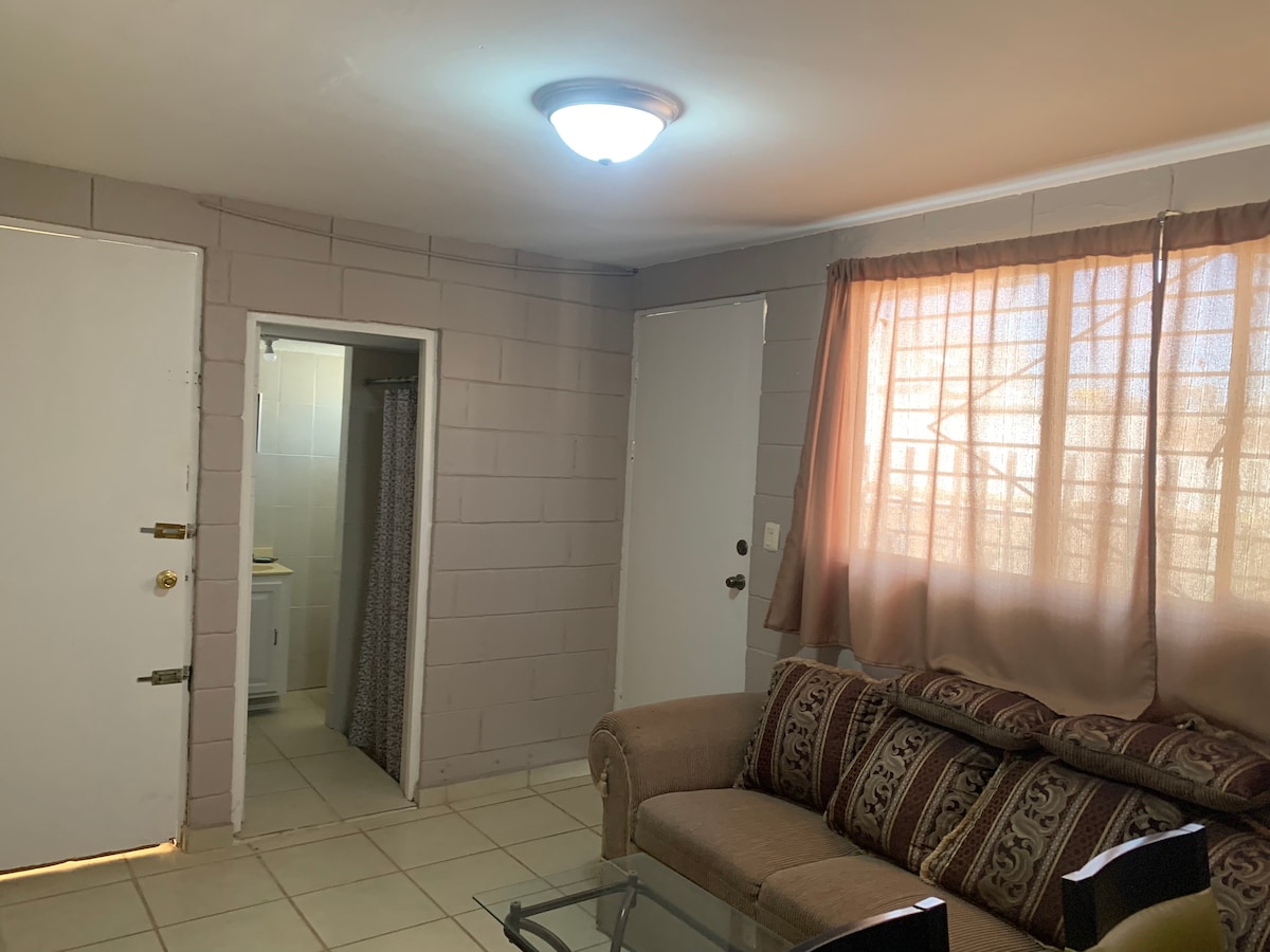 The living area features a comfortable sofa with decorative cushions, complemented by a glass coffee table. Natural light filters in through a window adorned with light curtains, illuminating the neutral-toned walls and tiled floor. A bathroom entrance is visible in the background.