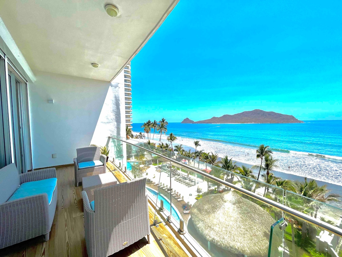 A private balcony features modern furniture with comfortable seating, offering unobstructed views of the ocean and beach. The clear blue sky complements the scenic landscape, while palm trees sway beside the shoreline, creating a serene outdoor space.
