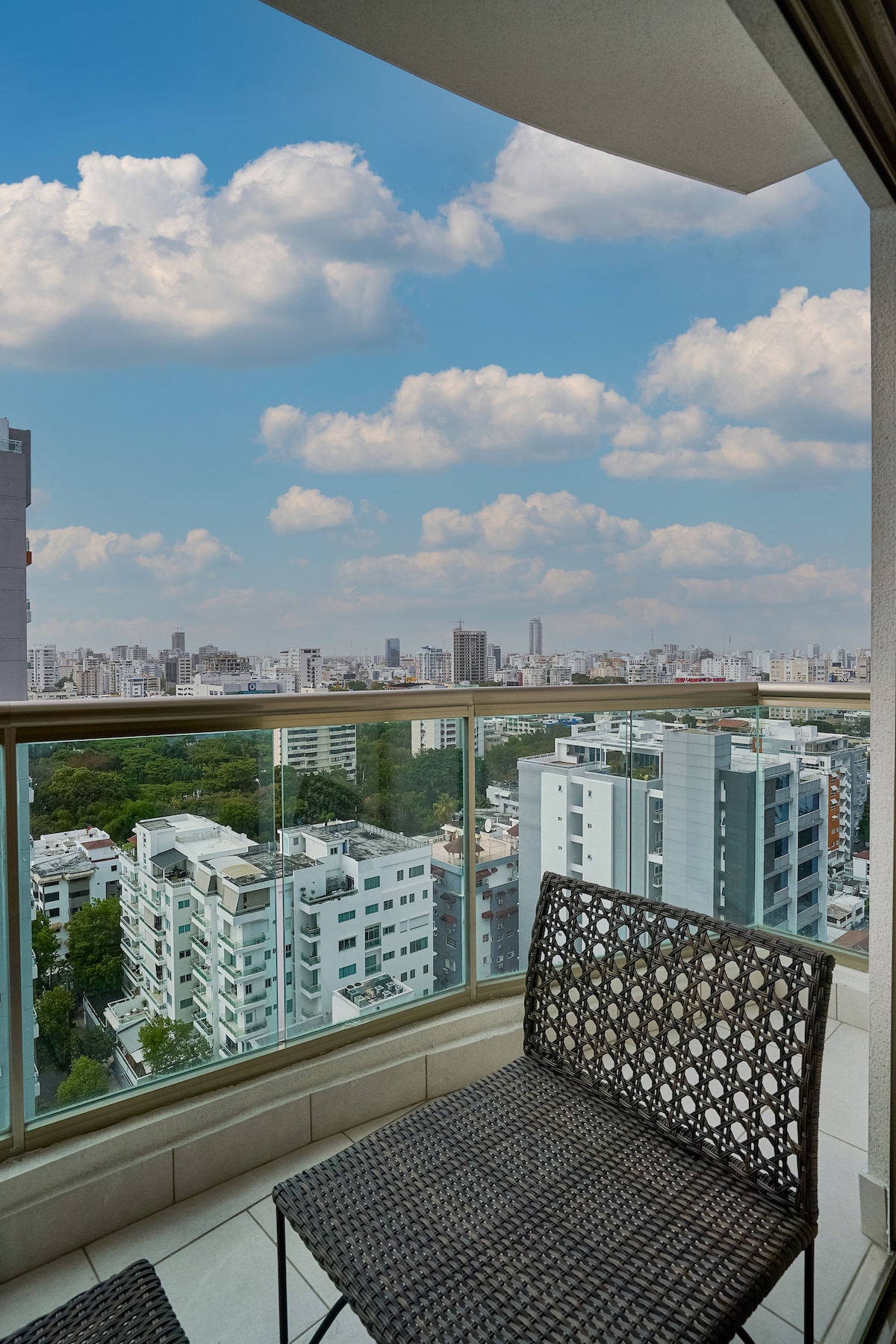 A balcony view showcases a vibrant urban landscape with a mix of modern buildings and green treetops. The glass railing provides an unobstructed view of the city skyline under a bright blue sky, dotted with fluffy white clouds. Two woven chairs are situated on the balcony.