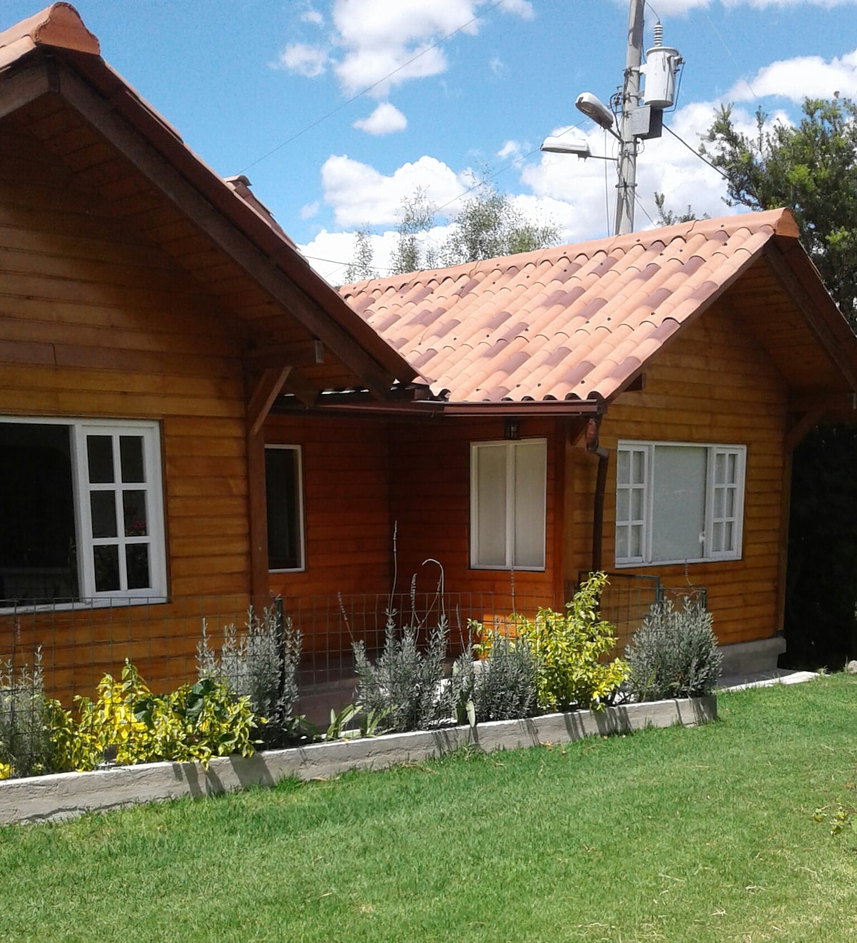 A wooden house features a charming exterior with a tiled roof and multiple windows. Lush green grass surrounds the property, complemented by vibrant flowers planted along the base. The building exudes a warm and inviting vibe, inviting relaxation in the natural setting.