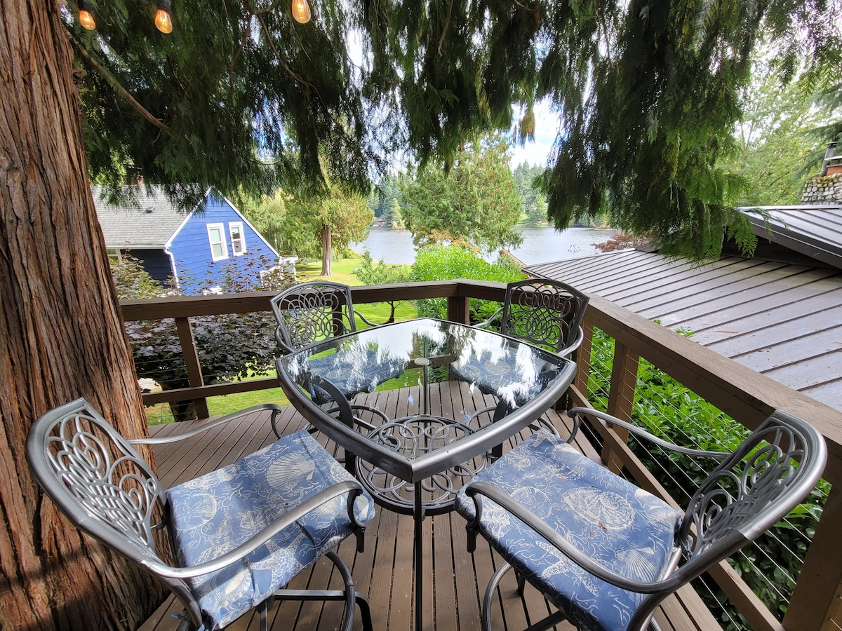 A deck table with four metal chairs sits under a canopy of trees, offering views of the lake beyond. The table is glass-topped, reflecting the surrounding greenery. Nearby buildings, including a blue house, complement the serene outdoor setting.