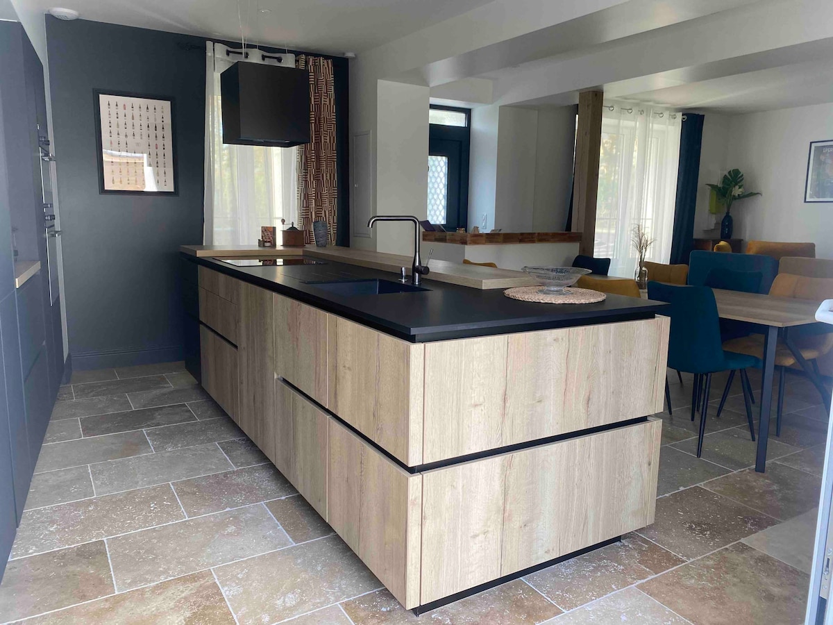A modern kitchen is presented with a central island featuring a dark countertop. Sleek cabinetry complements the neutral stone flooring. Natural light filters through large windows, highlighting a dining area furnished with various colorful chairs and a cozy sofa within the open layout.