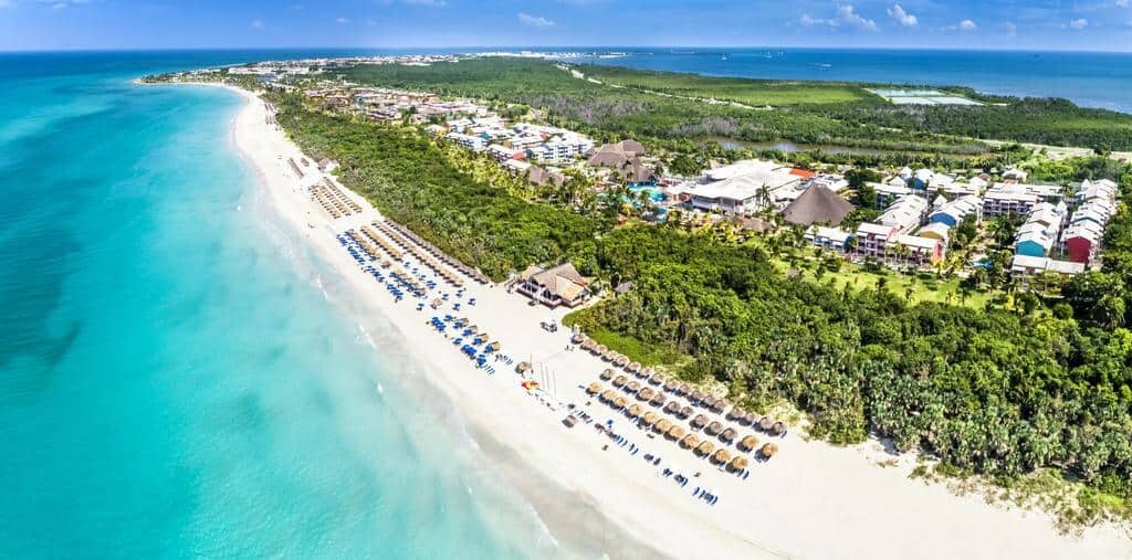 An aerial view displays a serene stretch of sandy beach alongside clear turquoise waters. Resort umbrellas and lounge chairs are arranged neatly on the shore, while lush greenery and colorful buildings are seen in the background, creating a harmonious coastal landscape.