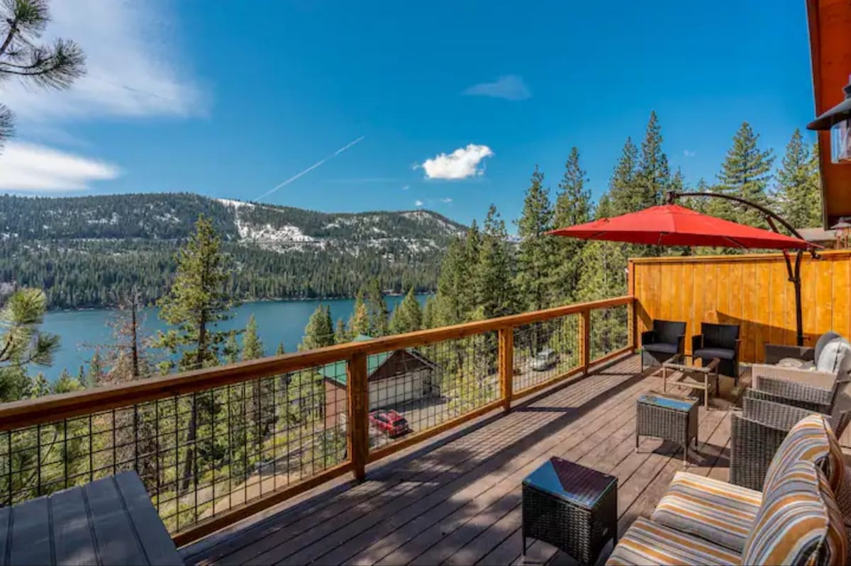 An expansive deck offers stunning panoramic views of Donner Lake and surrounding pine trees. Outdoor furniture, including four wicker chairs and a table, is arranged for comfort, complemented by a large red umbrella providing shade. The clear blue sky enhances the scenic tranquility.