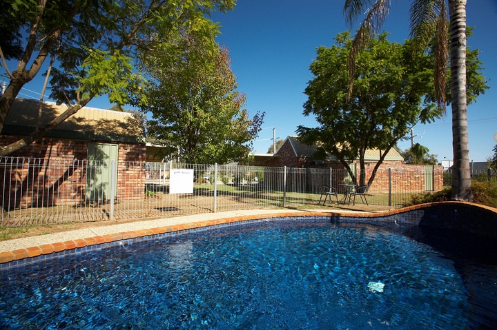 An inground saltwater pool is surrounded by a fenced area featuring lush greenery and mature trees. Nearby, a comfortable seating area with chairs is visible, adding to the relaxing atmosphere. The clear blue water of the pool reflects the bright sky above.