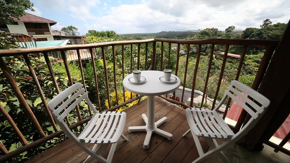 A private balcony presents a round table with two white chairs, overlooking lush greenery and rolling hills. Bright cups sit on the table, inviting a serene moment in nature, complemented by distant views of the Ecolodge and sky.