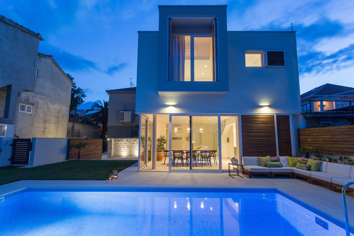 The modern villa is showcased at twilight, featuring large glass doors that open to a spacious interior. A crystal-clear swimming pool reflects the evening sky, and a comfortable outdoor seating area is positioned beside the pool, surrounded by a well-kept lawn.