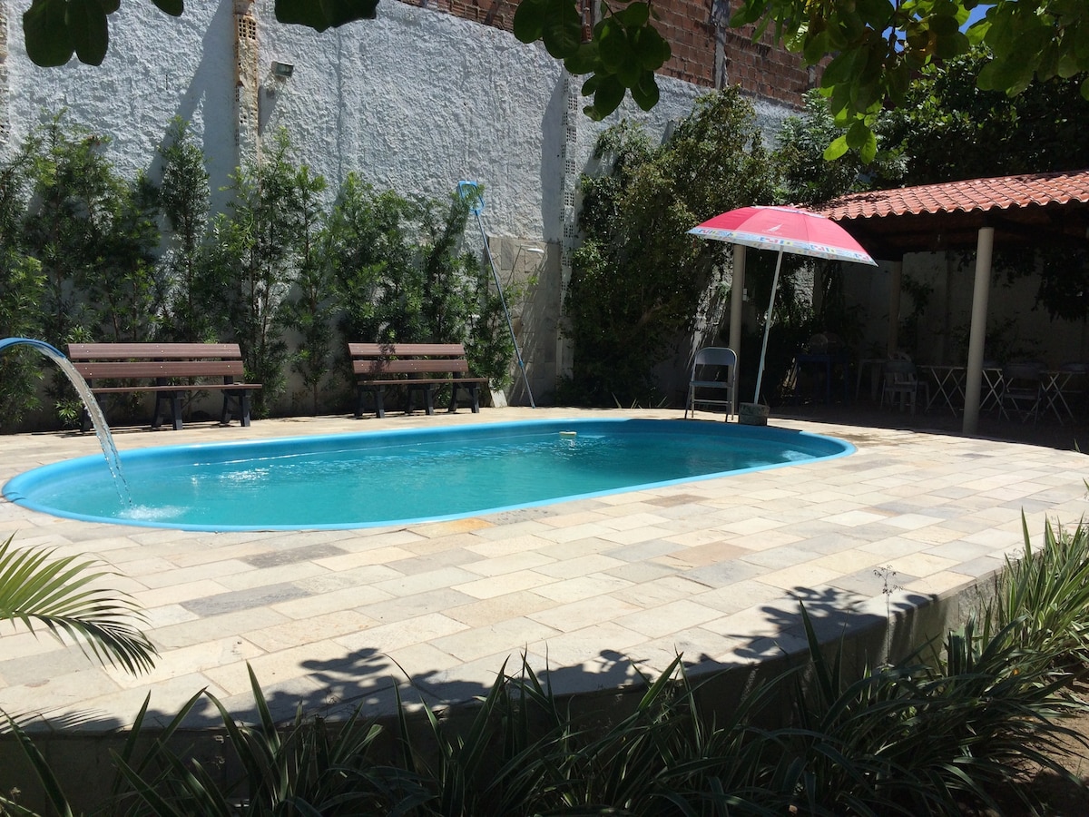 A private pool is surrounded by stone tiles, showcasing a clear blue water feature. Benches are positioned nearby under leafy greenery, while a shaded area with a parasol is visible to one side. Lush plants frame the scene, enhancing the tranquil environment.