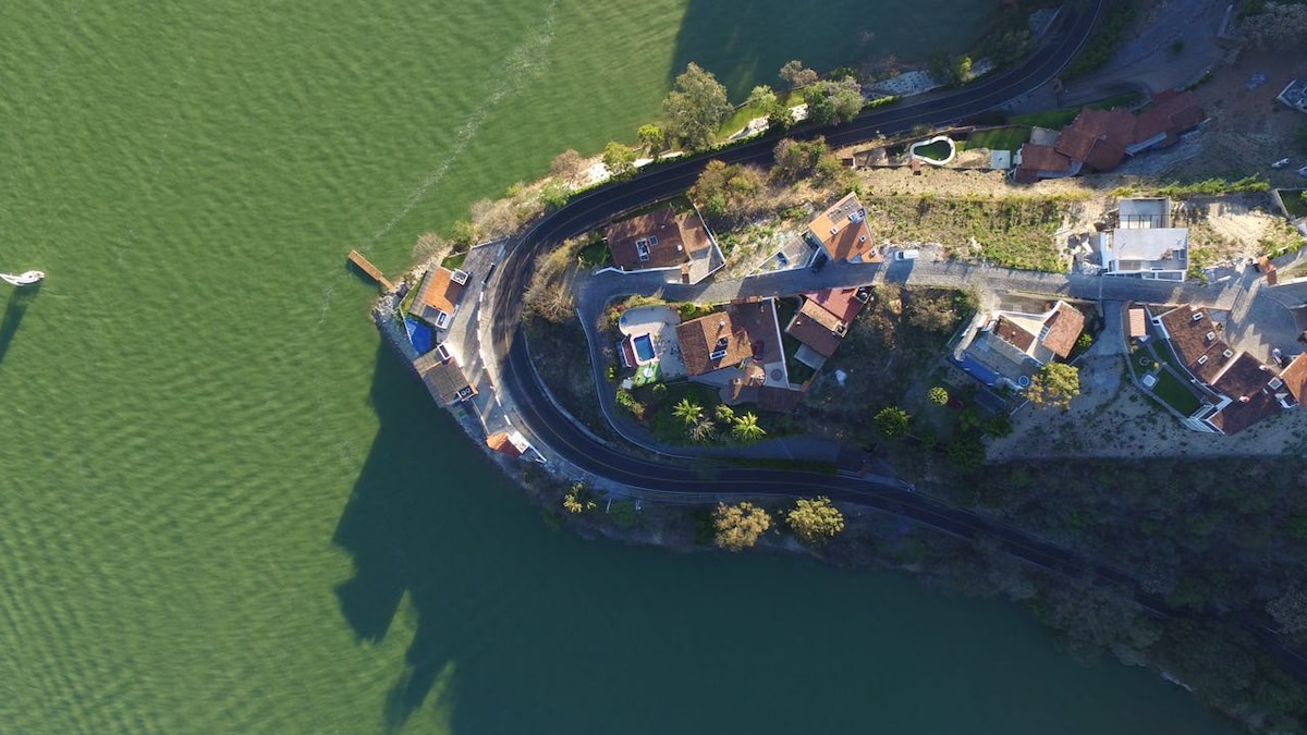An aerial view captures a scenic landscape featuring a collection of houses with red-tiled roofs lining the shoreline. A curved road surrounds the properties, while a patch of greenery separates them from the tranquil water.
