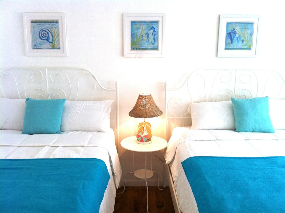 A bright sleeping area features two Queen beds adorned with white linens and aqua blue accents. A small bedside table with a woven lamp is placed between the beds, complemented by framed beach-themed artwork on the walls, enhancing the overall decor.