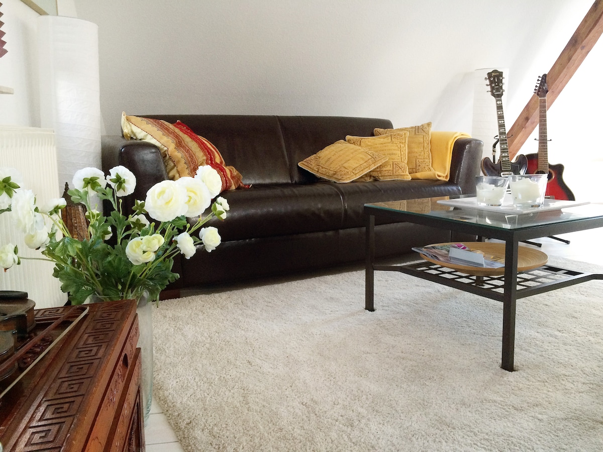 A cozy living area is showcased, featuring a dark sofa with decorative pillows and a glass coffee table. A floral arrangement sits nearby, complemented by a textured rug. Two guitars are displayed, adding a personal touch to the space.