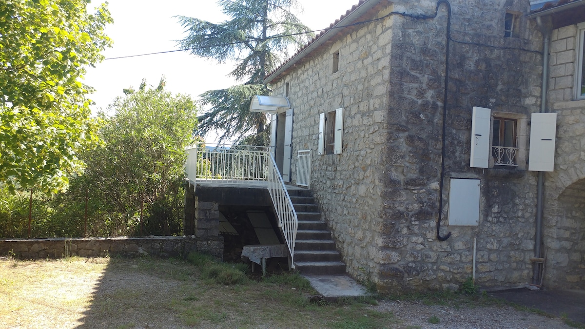 The exterior of a stone building is shown, featuring a staircase leading to a covered entrance. White shutters frame the windows, and lush greenery is visible in the surrounding area. An open space for outdoor activities is present in the foreground.