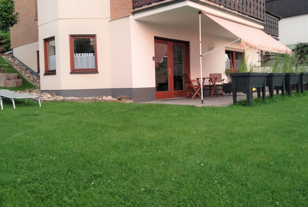 The image showcases a ground-level apartment with a patio area. Wooden doors lead to an inviting outdoor space, complete with a small table and chairs under an awning. A well-maintained grassy lawn surrounds the apartment, providing a natural setting.